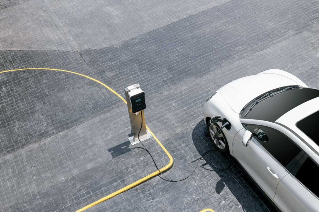 Overhead view of an electric vehicle connected to a charging station, with a yellow charging cable extending across a gray paved surface.